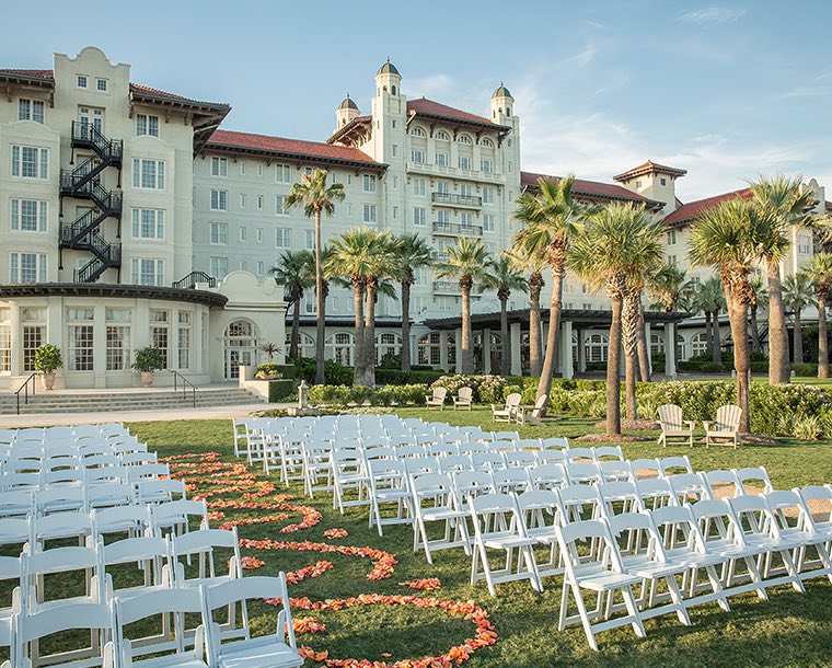 A wedding ceremony set up on the lawn outside Hotel Galvez with rows of white folding chairs and decorative flowers down the isle